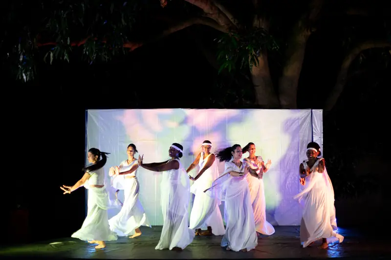 Huit danseuses en blanc devant un écran éclairé, sous un arbre, la nuit